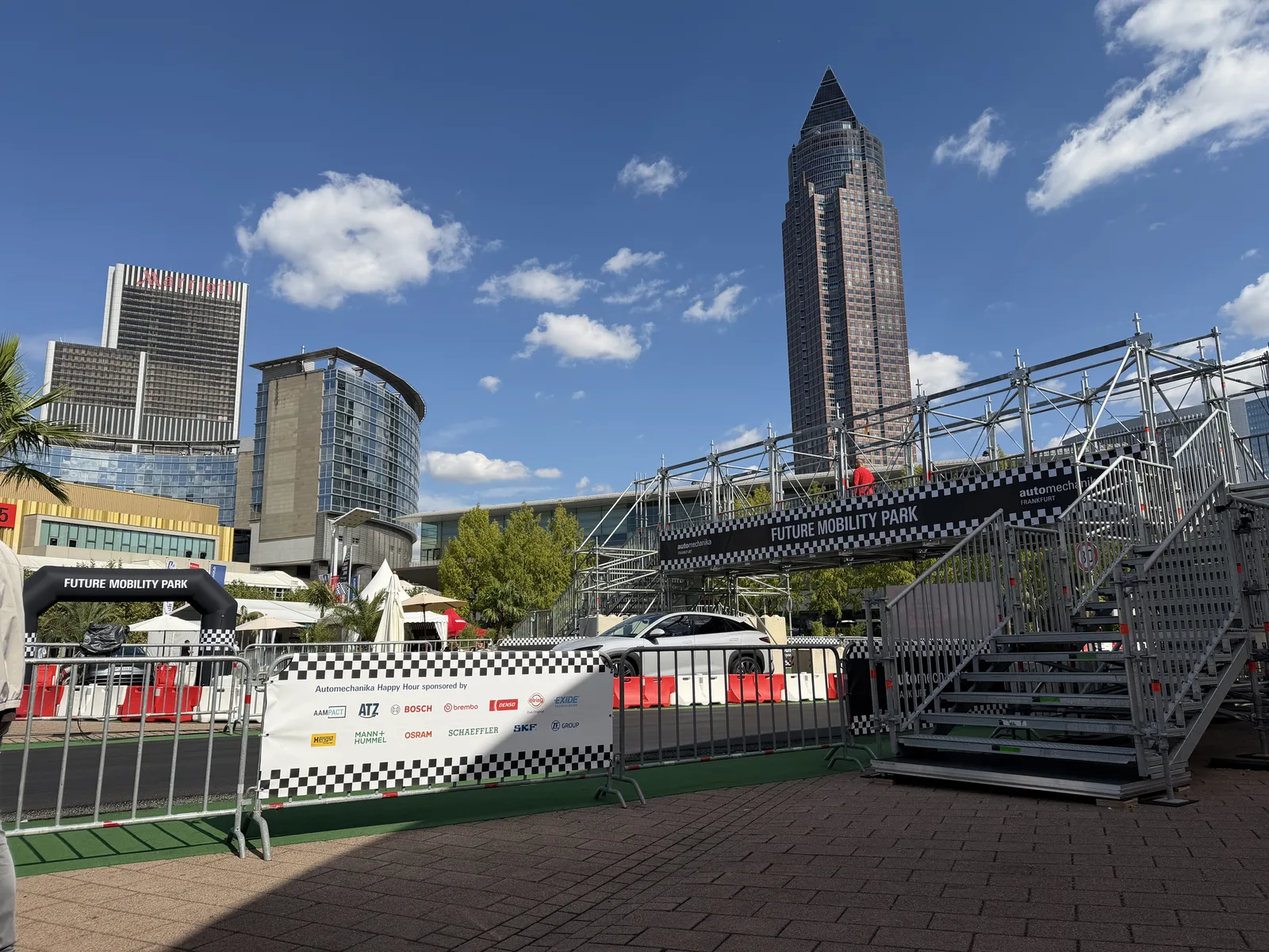 Wieża Messeturm i łuk strefy Future Mobility Park na Automechanika Frankfurt 2026, na tle błękitnego nieba